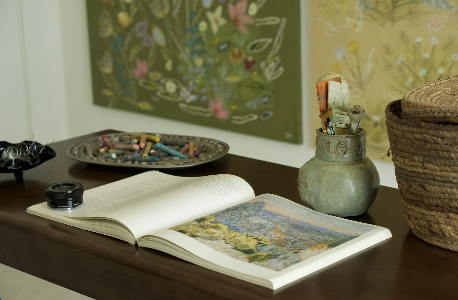 The studio table — a dish of worn pastels, an open art book and brushes in a stoneware jar